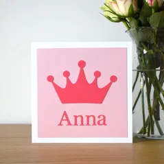 A photograph of a square card on a shelf next to a vase of flowers. The card is white with a light pink background and a darker pink crown shape on the front and a name underneath the crown.