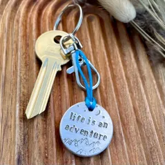 1 inch aluminium disc hand stamped with the words life is an adventure with moon and stars design above and mountains below.  shown on a royal blue cord turning the disc into a hanging keyring