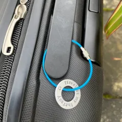 1 inch aluminium washer hand stamped around the disc with the words travel is my therapy in lowercase font with a small heart.  shown threaded onto an  electric blue wire which creates an approx 5cm loop as it attaches securely to handle of suitcase