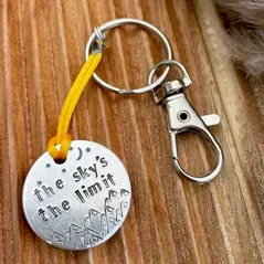 1 inch aluminium disc hand stamped with the words the sky's the limit with moon and stars design above and mountains below/  Shown on an orangy-yellow coloured cord turning the disc into a hanging keyring