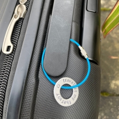 1 inch aluminium washer hand stamped around the disc with the words travel is my therapy in lowercase font with a small heart.  shown threaded onto an  electric blue wire which creates an approx 5cm loop as it attaches securely to handle of suitcase