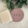 Image shows a round pink  jesmonite soap dish with 3 drainage holes, with a white soap bar resting on it, surrounded by eucalyptus whilst sat on a cream tile