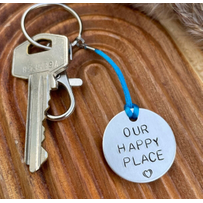 1 inch aluminium disc hand stamped with the words our happy place in capital letters with a small heart below.  shown on a royal blue cord to turn the disc into a hanging keyring