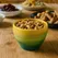 A handmade yellow and green ceramic bowl filled with mixed nuts, displayed on a rustic oak table in a farmhouse kitchen, surrounded by buffet-style food and warm wooden cabinetry.