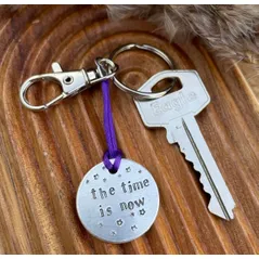 1 inch aluminium disc hand stamped with the words the time is now in lower case font, with small stars above and below.  shown on a purple cord to turn the disc into a hanging keyring