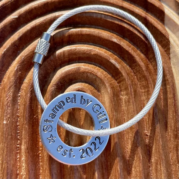 1 inch aluminium washer that can be personalised.  the image shows it stamped with the words stamped by gill est 2022.  the washer is threaded onto a silver wire loop to make a luggage tag.