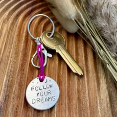 1 inch aluminium disc hand stamped with the words follow your dreams in capital letters, with small moon and stars design above.  Shown on a deep pink cord to turn the disc into a hanging keyring