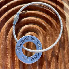 1 inch aluminium washer that can be personalised.  the image shows it stamped with the words stamped by gill est 2022.  the washer is threaded onto a silver wire loop to make a luggage tag.