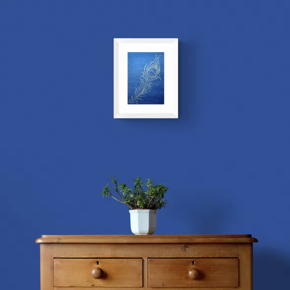 Bronze Peacock Feather on Navy Textured Paper Framed in White on a Blue Wall Above a Wooden Drawer Unit