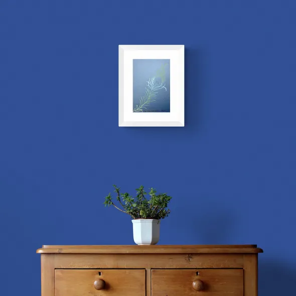 Ombre blue and green Peacock Feather on dusky blue Paper Framed in White on a Blue Wall Above a Wooden Drawer Unit