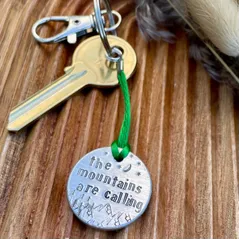 1 inch aluminium disc hand stamped with the words the mountains are calling with moon and stars design at the top and mountains at the bottom.  shown hung on a green cord turning it into a hanging keyring