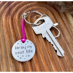 1 inch aluminium disc hand stamped with the words living my best life in lowercase font with small stars above and below.  shown on a bright pink cord to turn the disc into a hanging keyring