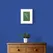 White Peacock Feather on Green Textured Paper Framed in White on a Blue Wall Above a Wooden Drawer Unit