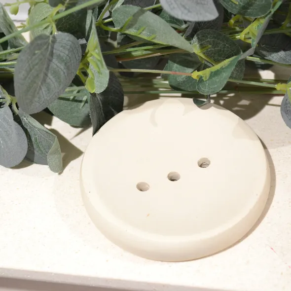 Image shows a cream coloured round soap dish with 3 drainage holes, on a cream background surrounded by eucalyptus
