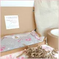 Open shipping box for a handmade item wrapped in white and pink tissue paper that is sealed with a sticker, a thank you note is stuck on the open flap of the box. Eco friendly packaging materials surround the box on a beige and white surface.