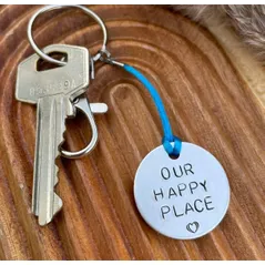 1 inch aluminium disc hand stamped with the words our happy place in capital letters with a small heart below.  shown on a royal blue cord to turn the disc into a hanging keyring