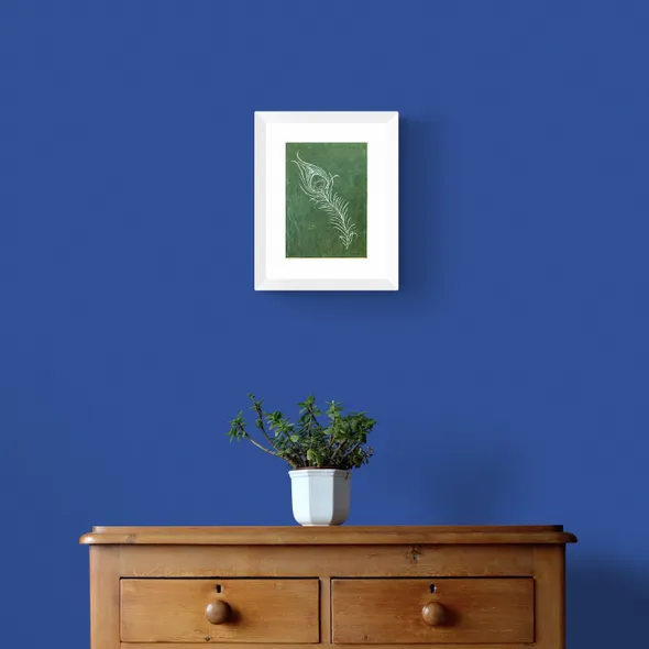 White Peacock Feather on Green Textured Paper Framed in White on a Blue Wall Above a Wooden Drawer Unit