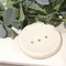 Image shows a cream coloured round soap dish with 3 drainage holes, on a cream background surrounded by eucalyptus