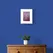 Bronze Peacock Feather on Burgundy Textured Paper Framed in White on a Blue Wall Above a Wooden Drawer Unit