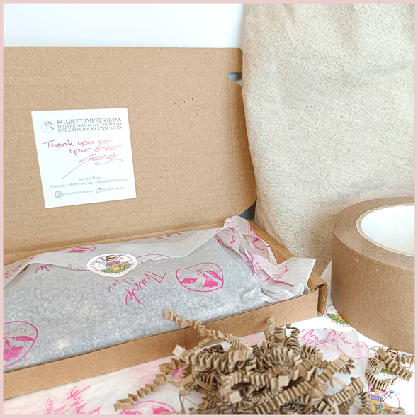 Open shipping box for a handmade item wrapped in white and pink tissue paper that is sealed with a sticker, a thank you note is stuck on the open flap of the box. Eco friendly packaging materials surround the box on a beige and white surface.