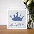 A photograph of a square card on a shelf next to a vase of flowers. The card is white with a light blue background and a darker blue crown shape on the front and a name underneath the crown.