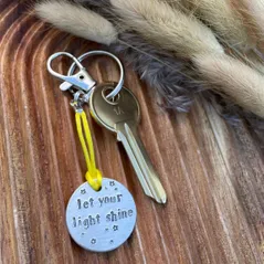 in inch aluminium disc hand stamped with the words let your light shine in lowercase font with small stars above and below.  Shown on a yellow cord to turn the disc into a hanging keyring