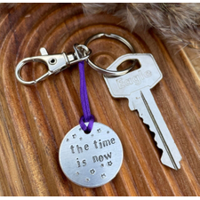 1 inch aluminium disc hand stamped with the words the time is now in lower case font, with small stars above and below.  shown on a purple cord to turn the disc into a hanging keyring