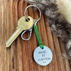 1 inch aluminium disc hand stamped with the words make it happen in capital letters with a small arrow stamp below.  shown on a green cord turning the disc into a hanging keyring