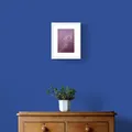 Bronze Peacock Feather on Burgundy Textured Paper Framed in White on a Blue Wall Above a Wooden Drawer Unit