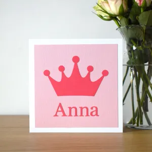 A photograph of a square card on a shelf next to a vase of flowers. The card is white with a light pink background and a darker pink crown shape on the front and a name underneath the crown.