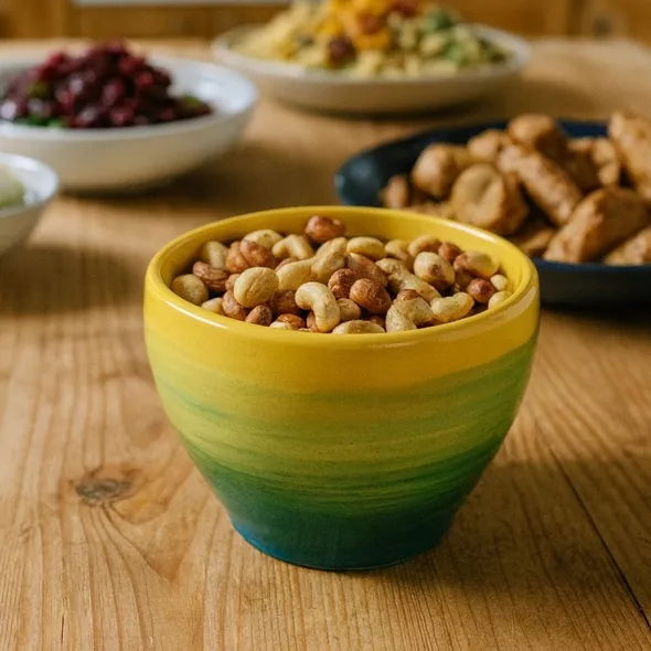 A handmade yellow and green ceramic bowl filled with mixed nuts, displayed on a rustic oak table in a farmhouse kitchen, surrounded by buffet-style food and warm wooden cabinetry.