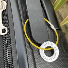1 inch aluminium washer stamped with the words life is an adventure.  the washer has been threaded onto a yellow gold wire to make a luggage tag.  the item is shown attached to the handle of a suitcase.