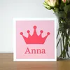 A photograph of a square card on a shelf next to a vase of flowers. The card is white with a light pink background and a darker pink crown shape on the front and a name underneath the crown.