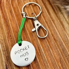 1 inch aluminium circular disc hand stamped with the words pocket hug in capital letters with a small heart below.  shown on a green coloured cord to make it into a hanging keyring