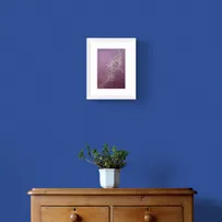 Bronze Peacock Feather on Burgundy Textured Paper Framed in White on a Blue Wall Above a Wooden Drawer Unit