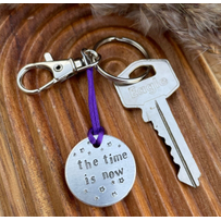 1 inch aluminium disc hand stamped with the words the time is now in lower case font, with small stars above and below.  shown on a purple cord to turn the disc into a hanging keyring