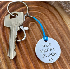 1 inch aluminium disc hand stamped with the words our happy place in capital letters with a small heart below.  shown on a royal blue cord to turn the disc into a hanging keyring