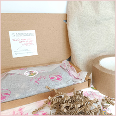 Open shipping box for a handmade item wrapped in white and pink tissue paper that is sealed with a sticker, a thank you note is stuck on the open flap of the box. Eco friendly packaging materials surround the box on a beige and white surface.